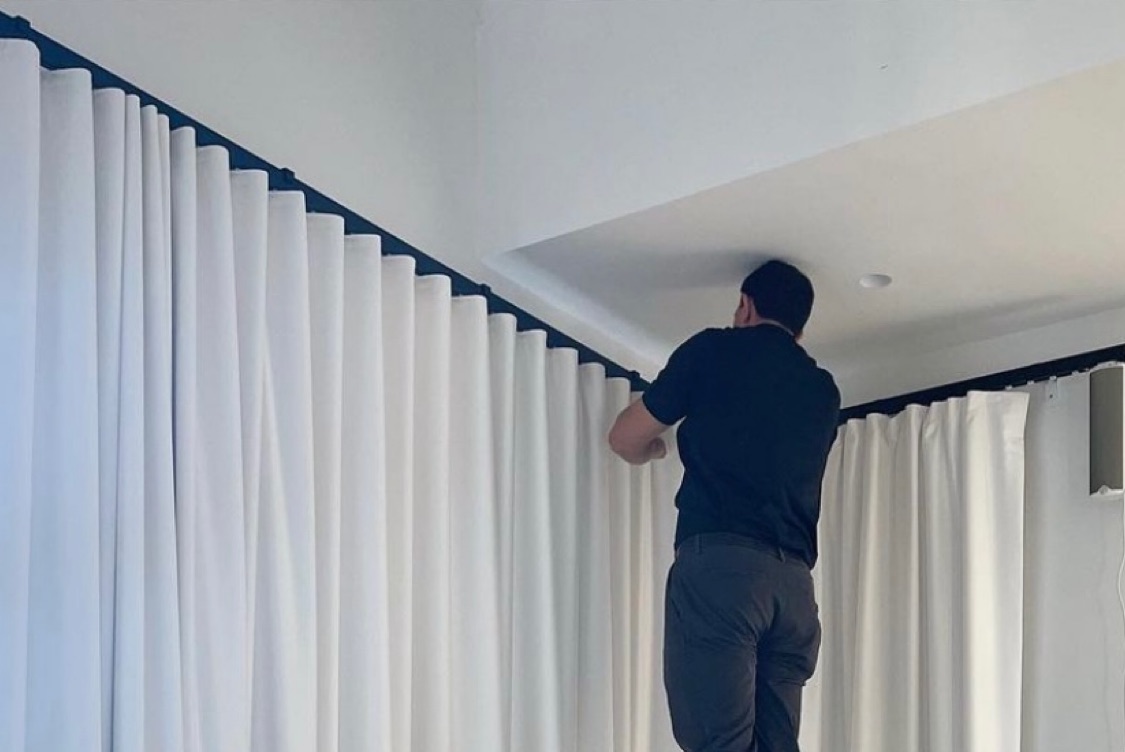 A professional installer putting up drapes inside a home. The drapes are predominately white.